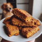 A stack of freshly baked Peanut Butter Pumpkin Baked Dog Treats on a plate, with a dog in the background.