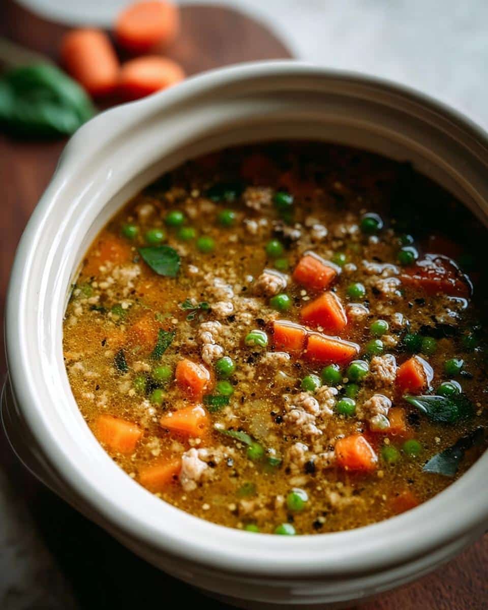 Close-up of a warm Carrot Pea Cozy Dog Broth Bowl filled with ground meat, bright green peas, and diced carrots in a savory broth.