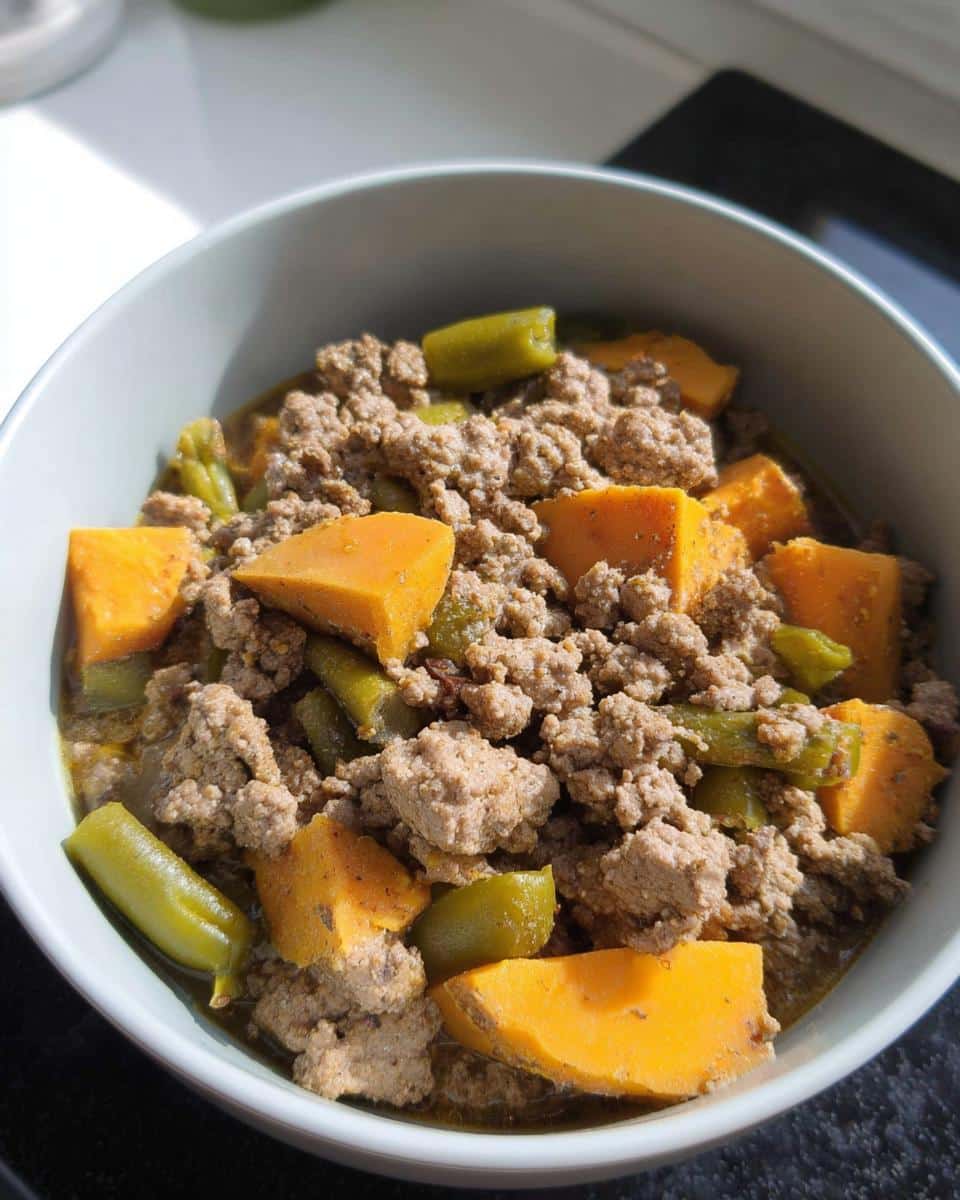 Close-up of Turkey-Free Holiday Dog Stew featuring ground meat, bright orange sweet potato chunks, and green beans in a light bowl.
