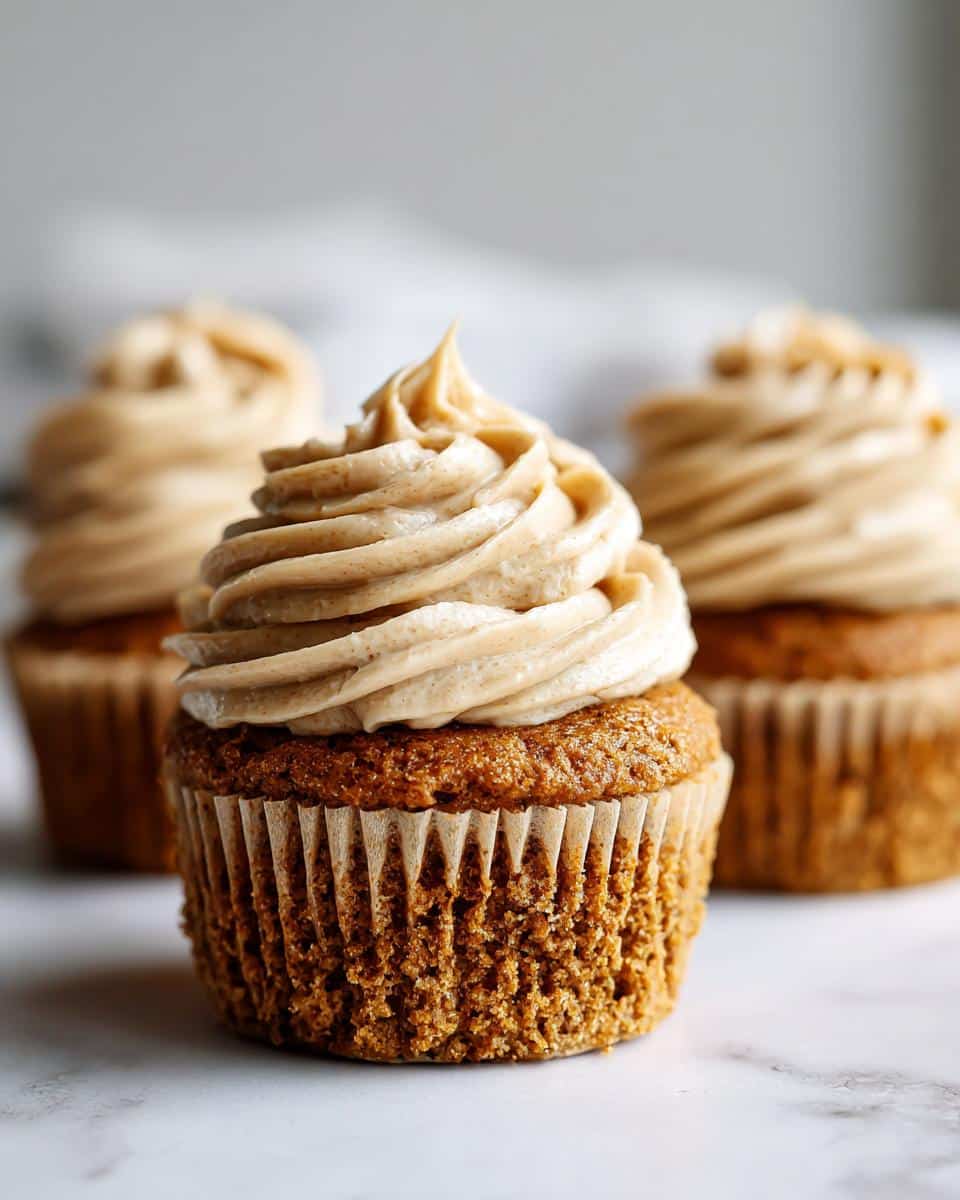 A close-up of one perfectly frosted Applesauce Pumpkin Pupcakes sitting on a white marble surface, with two others blurred in the background.