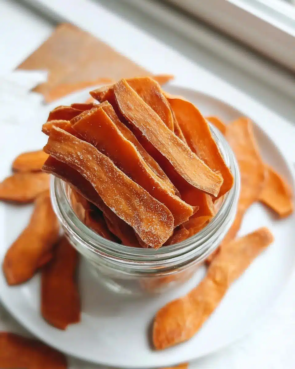 A glass jar filled with dehydrated orange strips of Fresh Sweet Potato Chews For Dogs, resting on a white plate.