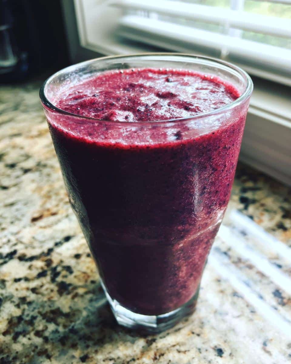 A tall glass filled with a thick, dark purple Frozen Blueberry Banana smoothie sitting on a granite countertop.