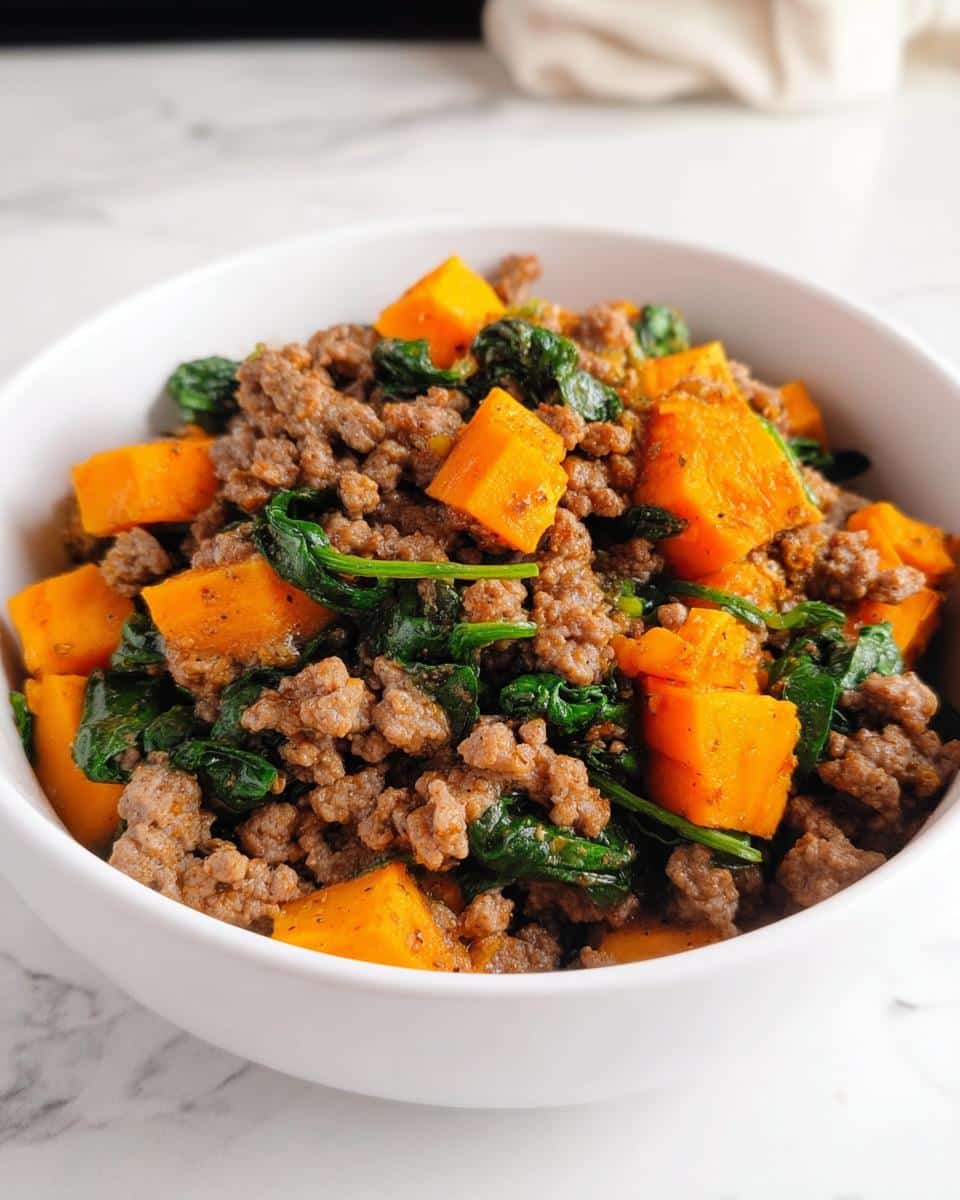 Close-up of the Beef and Sweet Potato Medley dog recipe mixture in a white bowl, featuring ground beef, orange sweet potato cubes, and wilted spinach.