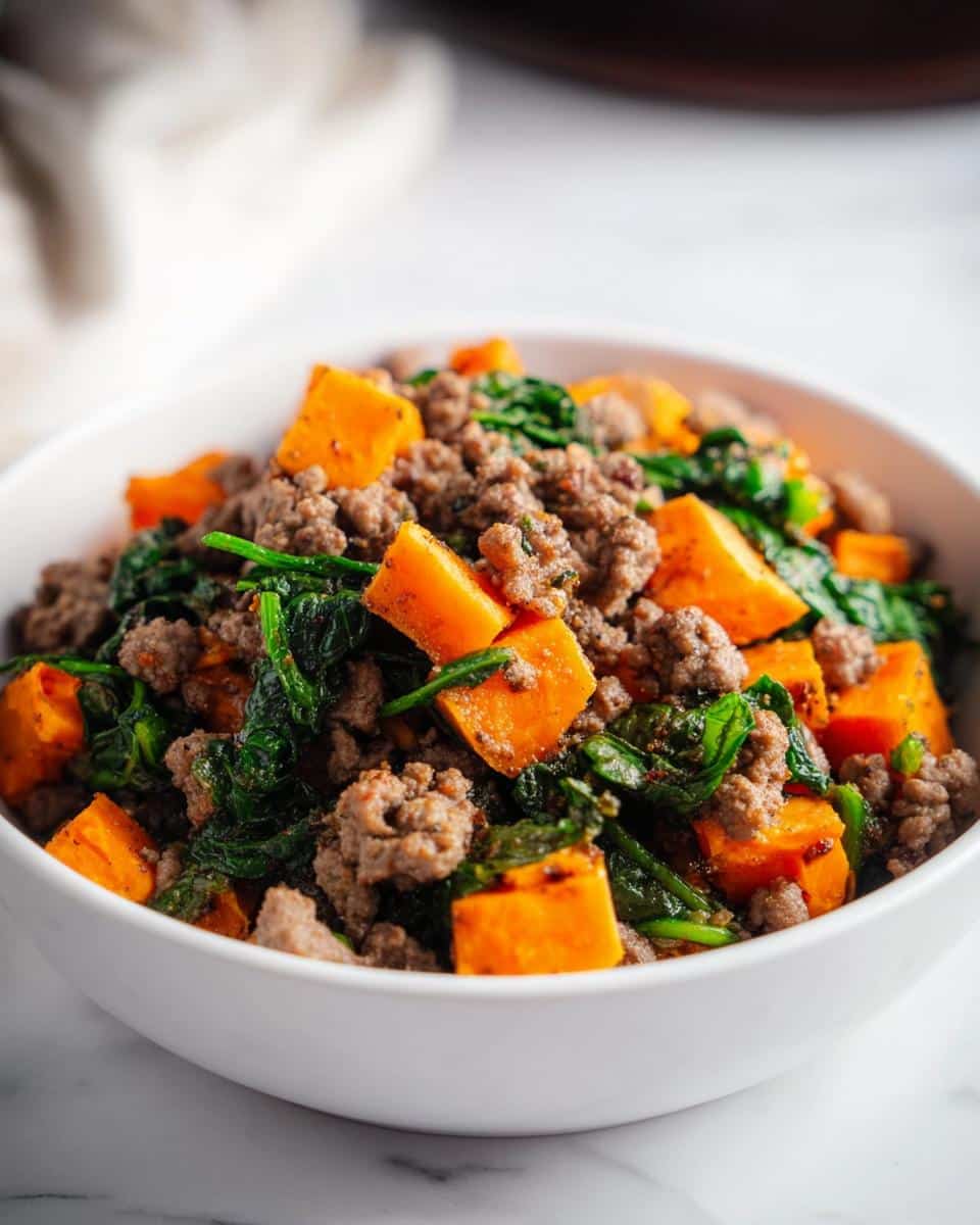 Close-up of a white bowl filled with the Beef and Sweet Potato Medley dog recipe, showing ground beef, orange sweet potato chunks, and dark green spinach.