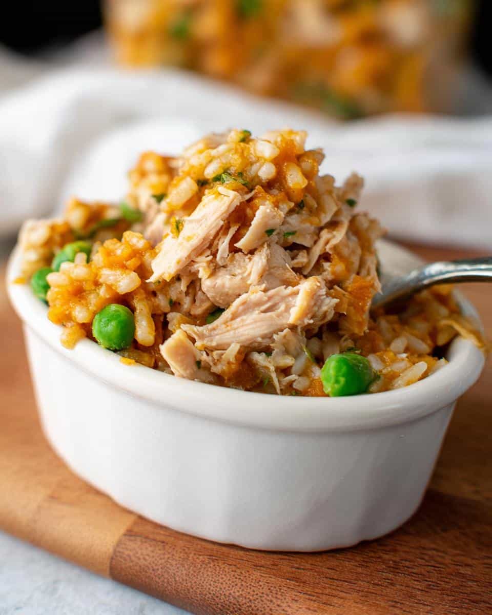 A close-up of a white bowl filled with homemade dog food: shredded chicken, pumpkin puree, white rice, and peas.